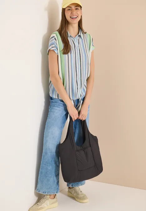 Frau trägt gestreiftes Hemd, weite Jeans, Sneaker und hält eine schwarze Tasche vor beigem Hintergrund.