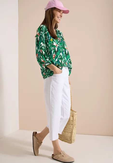 Eine Frau in grüner Blumenbluse, weißen Hosen und rosa Cap, mit Strohtasche und Mokassins.