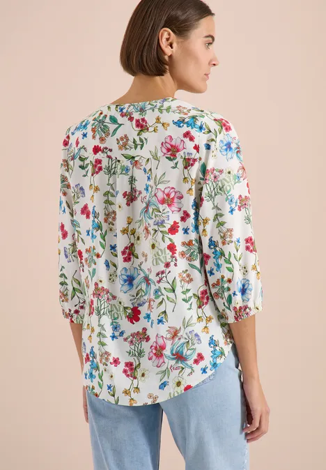 Femme vue de dos portant une chemise blanche à fleurs colorées et un jean, fond clair.
