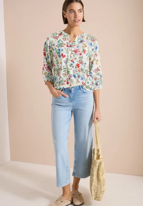 Femme portant un chemisier fleuri, jean bleu clair évasé, espadrilles et un sac en paille sur fond neutre.