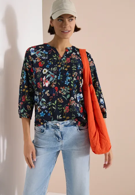 Chemisier à manches 3/4 avec col fendu et imprimé universal blue Une personne porte un chemisier bleu foncé à fleurs, un jean clair et un sac orange.