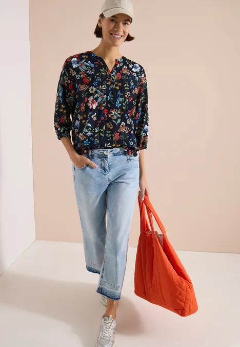 Chemisier à manches 3/4 avec col fendu et imprimé universal blue Femme en chemisier fleuri, jean bleu clair, baskets blanches et grand sac orange sur fond clair.