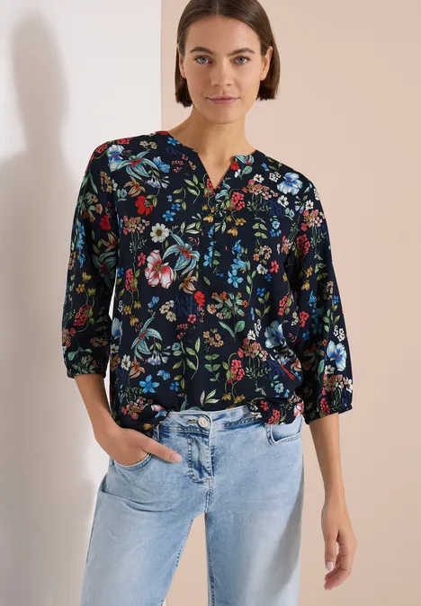 Chemisier à manches 3/4 avec col fendu et imprimé universal blue Femme portant un chemisier noir à fleurs colorées et manches 3/4 avec jean bleu clair.