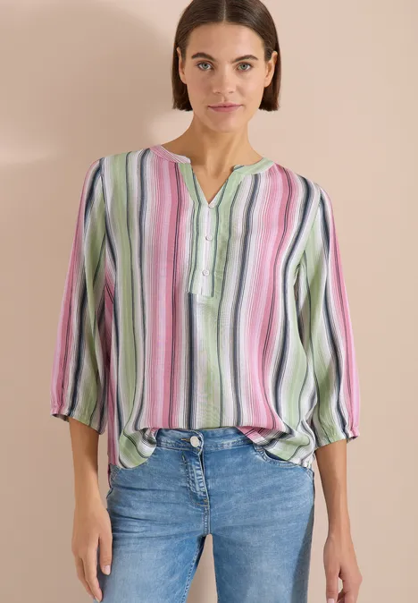 Frau in bunter, gestreifter Bluse und Jeans vor neutralem Hintergrund.