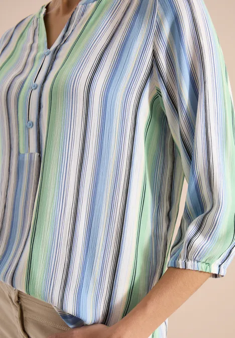 Chemisier à manches 3/4 avec rayures multicolores light sky blue Une personne porte un chemisier pastel rayé aux manches 3/4 retroussées, associé à un pantalon beige.