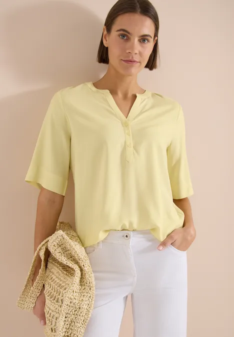 Frau in pastellgelber V-Bluse mit halblangen Ärmeln, weißer Hose und geflochtener Strohtasche vor hellem Hintergrund