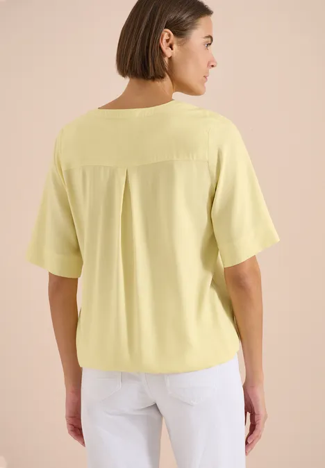 Frau von hinten in hellem Gelbshirt mit kurzen Ärmeln und weißer Hose.