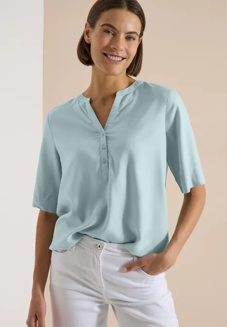 Kurzarm Bluse mit Split Neck crystal mint Eine Frau in einer hellblauen Bluse mit halblangen Ärmeln und weißen Hosen, eine Hand in der Tasche.