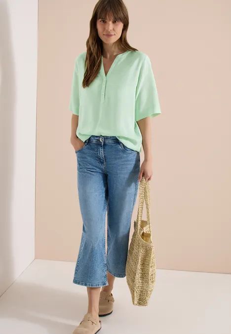 Frau in pastellgrünem T-Shirt, weitem Jeans-Culotte und Basttasche vor schlichtem Hintergrund.