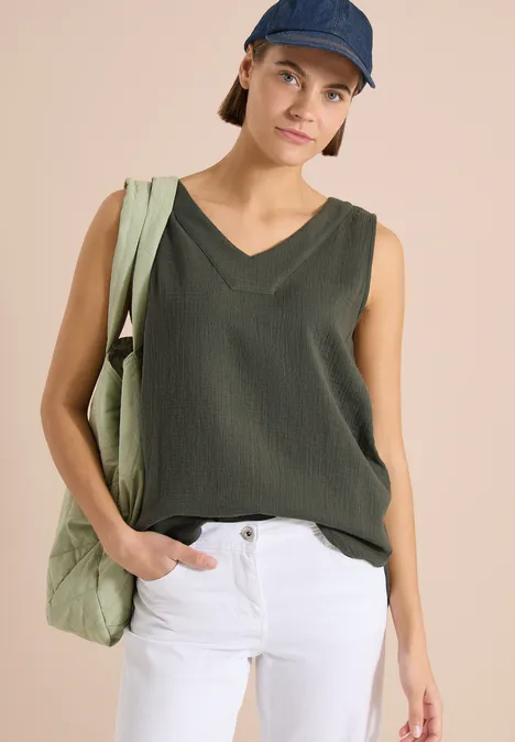 Vrouw in olijfgroene mouwloze top, witte broek en pet, met een stoffen tas nonchalant.