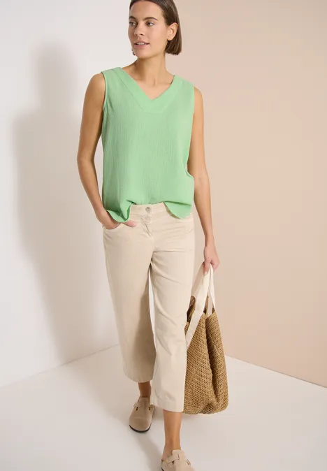 Vrouw met mouwloze mintgroene top, crèmekleurige broek tot op de kuit, beige instappers en strooien tas voor neutrale achtergrond.
