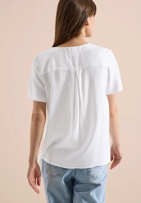 Frau von hinten in weißer, kurzärmliger Bluse und hellen Jeans vor beigem Hintergrund.