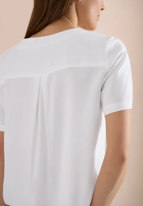 Eine Frau von hinten in weißer Bluse mit Rückendetail und kurzen Ärmeln vor neutralem Hintergrund.