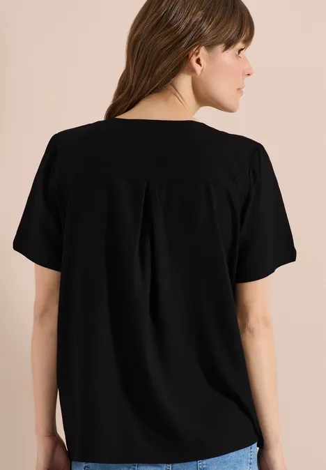 Rückenansicht einer Frau in einem schlichten, schwarzen Kurzarmshirt vor neutralem Hintergrund.