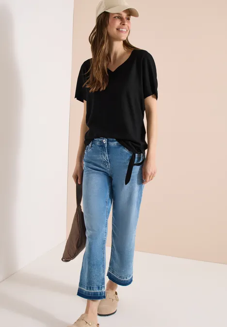 Frau trägt schwarzes T-Shirt, weit geschnittene hellblaue Jeans, beige Schuhe und hält eine taupefarbene Jacke.