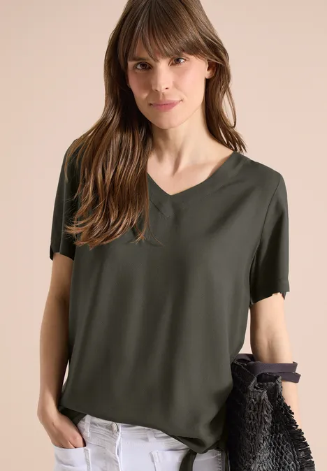 Frau trägt olivgrünes T-Shirt mit V-Ausschnitt, weiße Hose und hält eine schwarze Flecht-Tasche vor beigem Hintergrund.