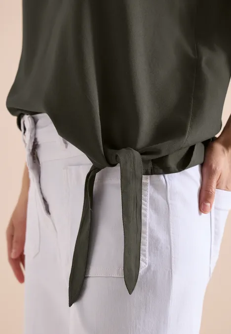 Eine Person in hellen Jeans trägt ein olivgrünes Oberteil, das seitlich geknotet ist.