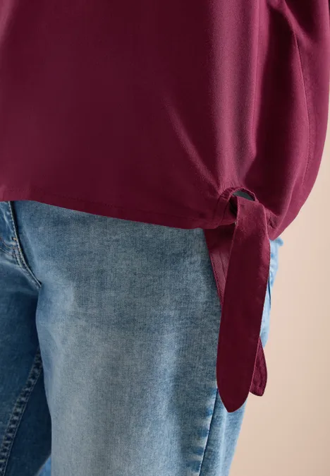 Hüftnahe Aufnahme: Jeans und ein seitlich geknotetes, burgunderrotes Oberteil.