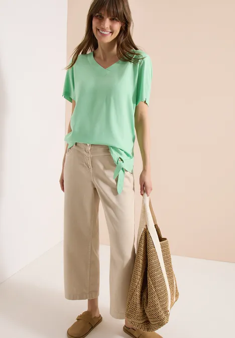 Modell mit mintgrünem V-Shirt, beigen Culottes und großer Strohtasche in schlichter Studio-Umgebung.