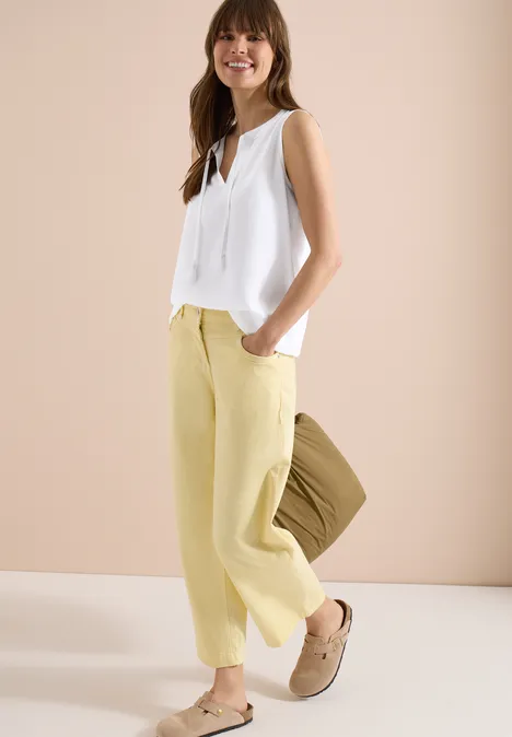 Frau vor hellem Hintergrund mit weißer ärmelloser Bluse, pastellgelber Hose, beigen Clogs und brauner Tasche.