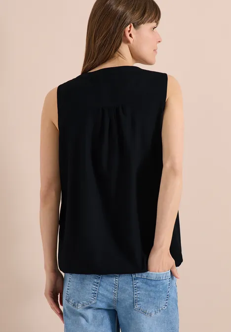 Frau von hinten in schwarzem ärmellosem Oberteil und blauer Jeans, Hände in den Taschen.