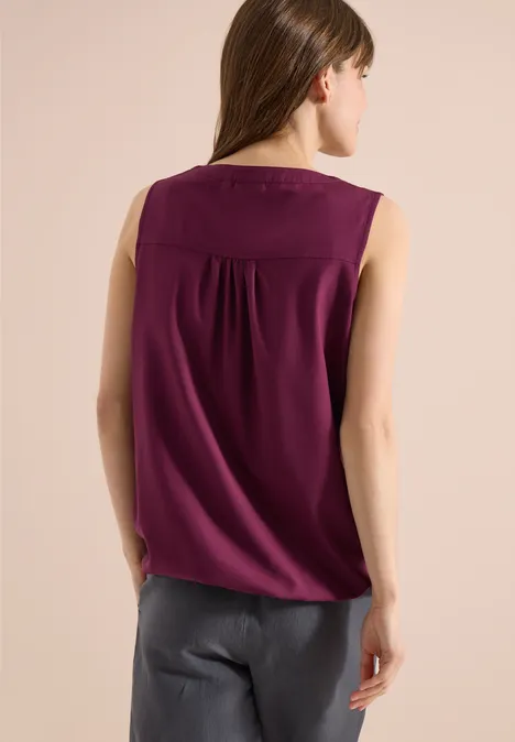 Frau von hinten in ärmellosem lila Top und grauer Hose vor hellem Hintergrund.