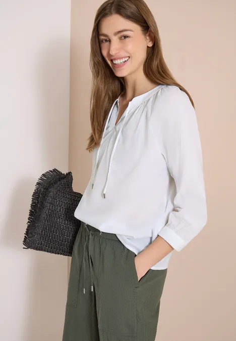 Vrouw in witte blouse en olijfgroene broek, met zwarte gevlochten clutch, voor een beige achtergrond.