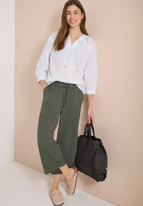 Vrouw in wit overhemd, losse groene broek, beige slippers en zwarte gevlochten tas.