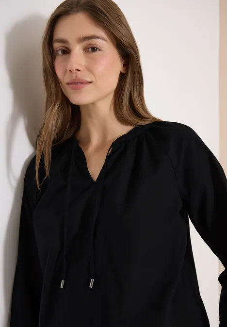 Eine Frau in einer schwarzen Bluse lehnt lässig an der Wand.