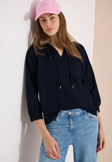 Eine Frau lehnt an eine Wand, trägt eine dunkelblaue Bluse mit Kapuzenkordel und helle Jeans.