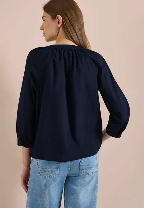 Eine Frau von hinten in einer marineblauen Bluse mit 3/4-Ärmeln und hellblauen Jeans vor beigem Hintergrund.