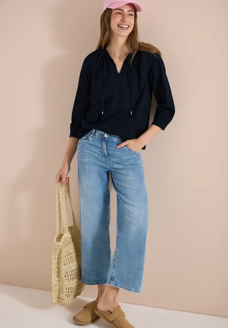 Frau in schwarzer Bluse, weiten Jeans und Basttasche steht vor beiger Wand.