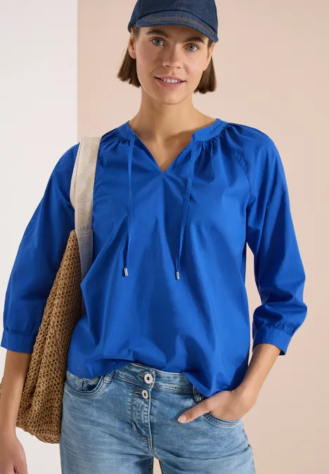 Person trägt blaue Bluse mit Kordeln, Jeans und Tasche vor neutralem Hintergrund.