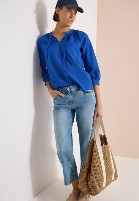 Frau lehnt an Wand mit blauer Bluse, hellen Jeans, Cap und großem geflochtenem Shopper.