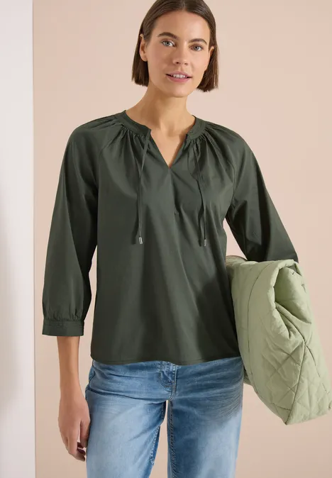 Frau trägt olivgrüne Bluse mit Kordelzug, Jeans und hält eine hellgrüne Steppjacke.