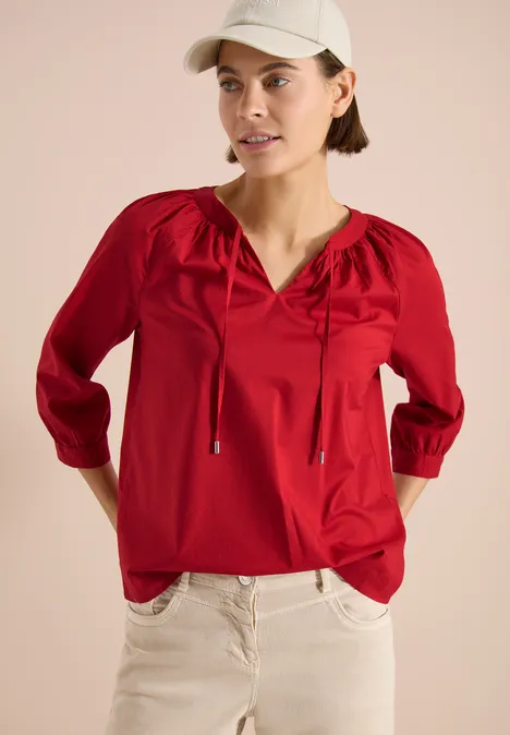 Person in roter Bluse mit 3/4-Ärmeln und Bändchen, kombiniert mit heller Hose und Cap vor beigem Hintergrund.