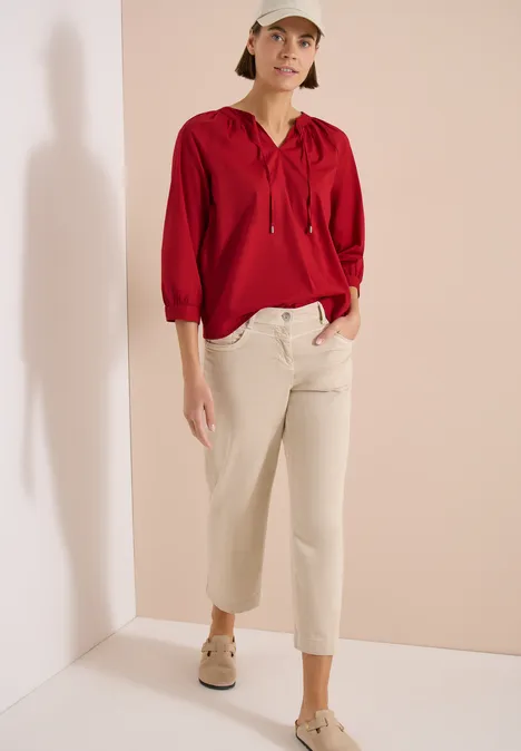 Frau in roter Bluse mit Kapuze, hellen Culotte-Jeans und beigen Schuhen vor hellem Hintergrund.