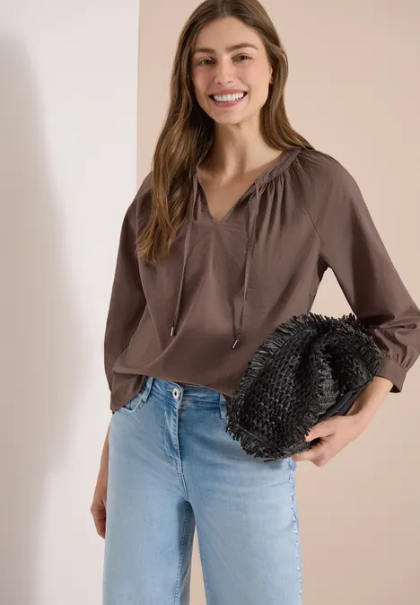 Vrouw in bruin shirt en spijkerbroek houdt zwarte clutch vast voor een lichte achtergrond.
