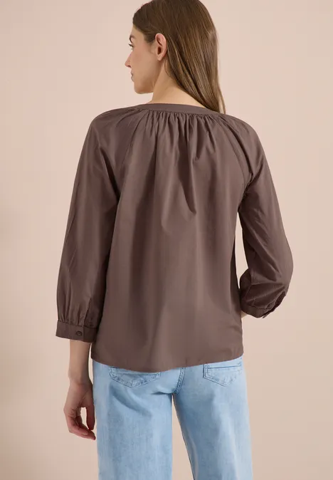 Vrouw van achteren in een bruine blouse met plooien en lichtblauwe spijkerbroek.