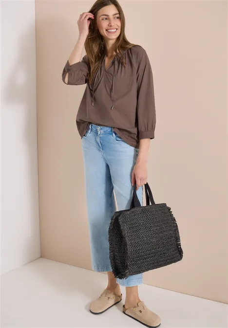 Vrouw in bruin shirt, lichte jeans en beige schoenen, met een grote zwarte gevlochten tas.