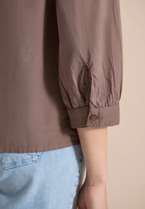 Een arm in een taupekleurig shirt met opgerolde mouw, gecombineerd met lichtblauwe jeans voor een neutrale achtergrond.