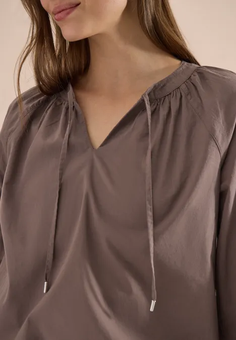 Close-up van een vrouw in een taupekleurige satijnen blouse met V-hals en siertouwtjes.
