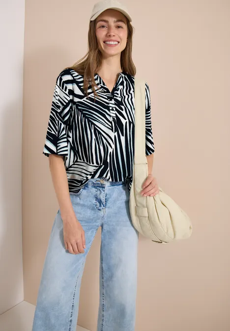 Frau in schwarz-weiß gestreifter Bluse, weiter hellblauer Jeans und beiger Schultertasche.