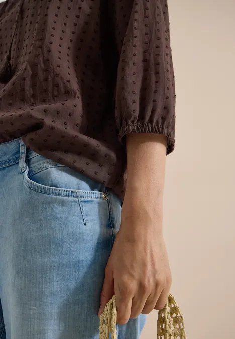 Eine Person in hellblauen Jeans und brauner Bluse hält eine beige, perlenbesetzte Handtasche.