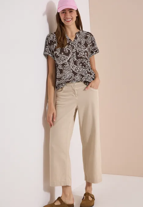 Blouse met split in de hals en print brown cacao Vrouw leunt tegen tweekleurige achtergrond, draagt een blouse met print, wijde beige broek en slingback-pumps.