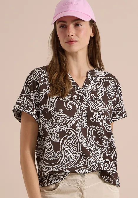 Blouse met split in de hals en print brown cacao Vrouw in een bruin t-shirt met wit paisleymotief, korte mouwen en V-hals voor een neutrale achtergrond.