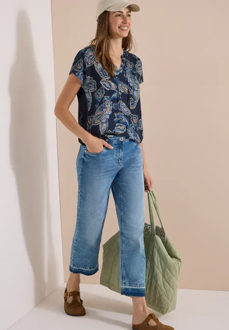 Blouse met split in de hals en een mix van texturen universal blue Vrouw met beige pet, gekleurde korte blouse, wijd korte spijkerbroek en grote groene gewatteerde tas.