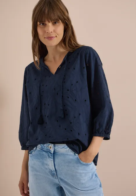 Frau vor beigem Hintergrund trägt eine locker sitzende dunkelblaue Lochstickerei-Bluse mit 3/4-Ärmeln und helle Jeans.