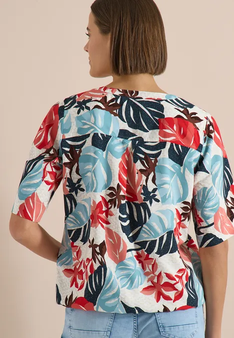 Rückenansicht einer Frau mit Kurzhaarschnitt, die eine kurzärmelige Bluse mit rot-blauem tropischem Blatt- und Blumenmuster trägt.