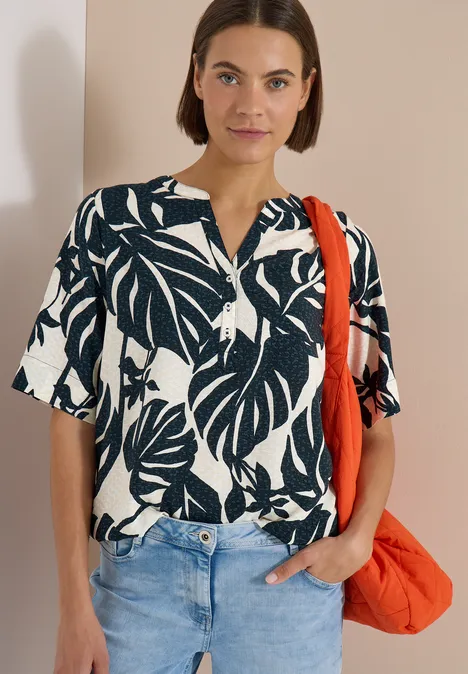Frau mit schwarz-weißem Blusen-Shirt mit Blattmuster, Jeans und orangefarbener Tasche vor beigem Hintergrund.